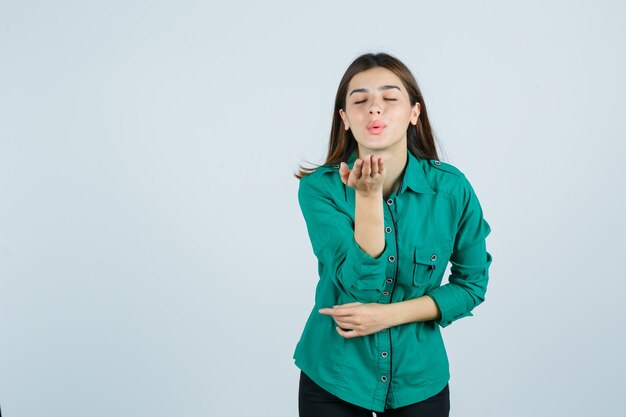 Hermosa joven enviando beso al aire con labios fruncidos en camisa verde y mirando pacífica, vista frontal.