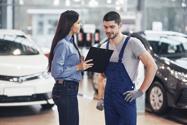 Hermosa joven está hablando con un guapo mecánico mientras repara un automóvil en concesionario