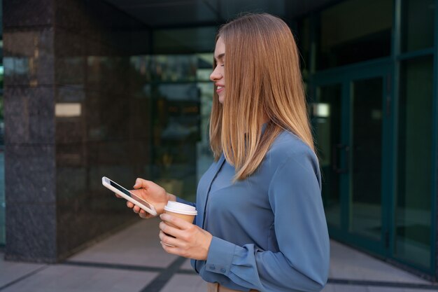 Hermosa joven está usando una aplicación en su dispositivo de teléfono inteligente para enviar un mensaje de texto cerca de edificios comerciales