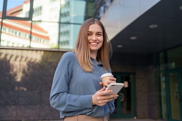 Hermosa joven está usando una aplicación en su dispositivo de teléfono inteligente para enviar un mensaje de texto cerca de edificios comerciales