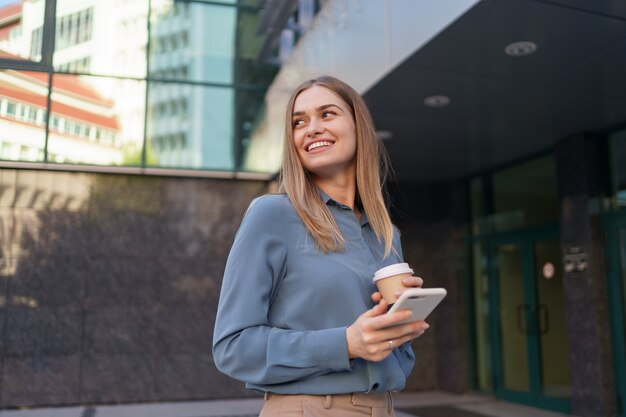 Hermosa joven está usando una aplicación en su dispositivo de teléfono inteligente para enviar un mensaje de texto cerca de edificios comerciales