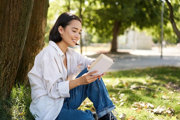 Hermosa joven estudiante asiática sentada en el parque bajo un árbol y leyendo un libro sonriendo disfrutando de su cálido