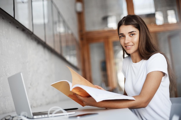 Hermosa joven estudiante preparándose para sus exámenes de inglés en el campus universitario usando una computadora portátil y un manual Concepto de educación