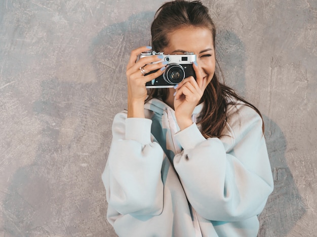 Hermosa joven fotógrafo sonriente chica tomando fotos con su cámara retro. Mujer haciendo fotos. Modelo vestida con sudadera casual de verano. Posando en estudio cerca de la pared gris