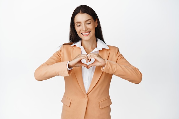 Hermosa joven gerente de oficina cierra los ojos y muestra el signo del corazón sonriendo despreocupado de pie en traje contra fondo blanco Copiar espacio