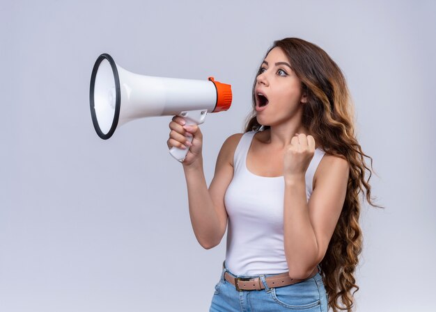 Hermosa joven gritando en altavoz mirando al lado izquierdo con el puño levantado en la pared blanca aislada