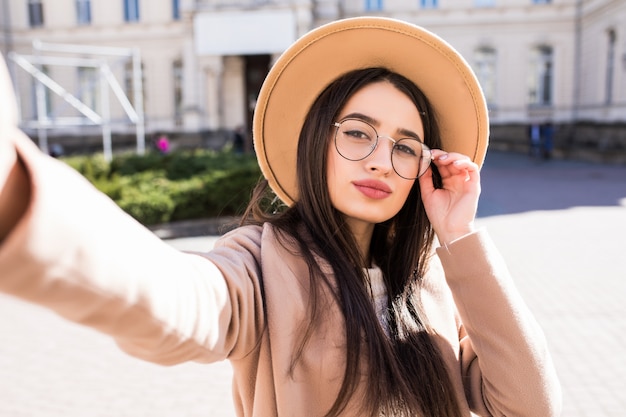 Hermosa joven hace selfie en su nuevo teléfono inteligente al aire libre en la ciudad en un día soleado