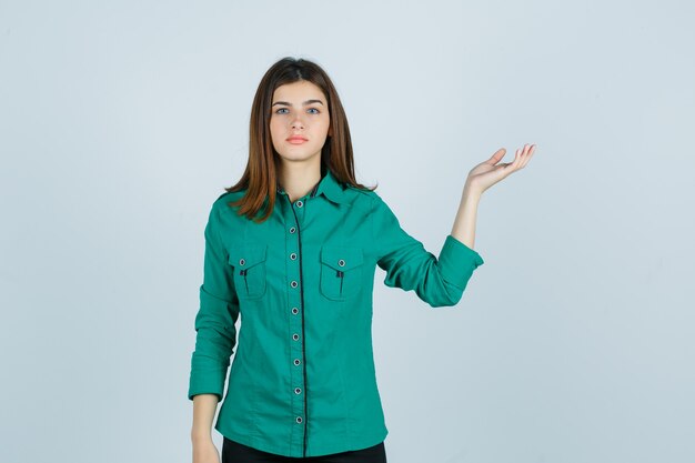 Hermosa joven haciendo gesto de bienvenida en camisa verde y mirando perplejo, vista frontal.