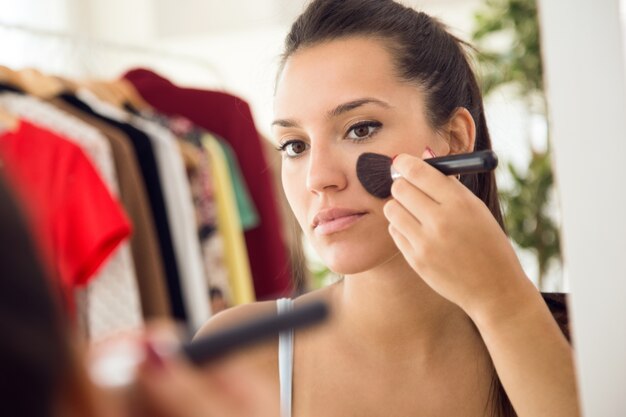 Hermosa joven haciendo maquillaje cerca de espejo en casa.