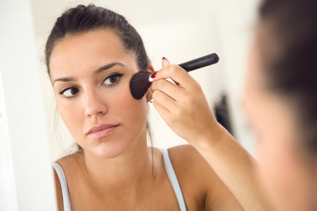 Hermosa joven haciendo maquillaje cerca de espejo en casa.