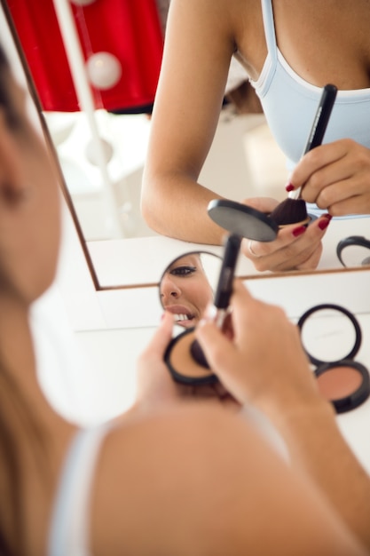 Hermosa joven haciendo maquillaje cerca de espejo en casa.