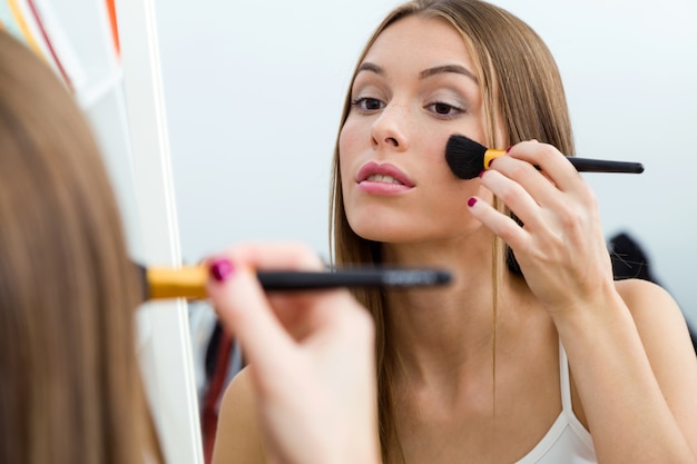 Hermosa joven haciendo maquillaje cerca de espejo en casa.