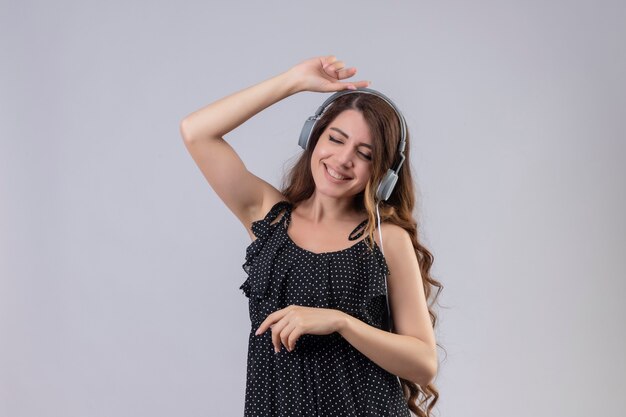 Hermosa joven inspirada en vestido de lunares disfrutando de su música favorita a través de auriculares inalámbricos divertidos bailando con los ojos cerrados sobre fondo blanco.