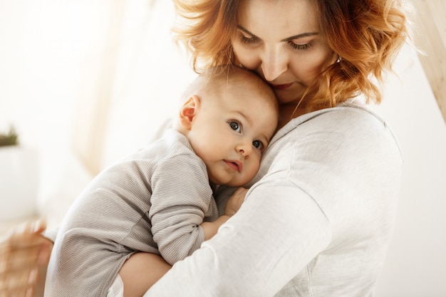 Hermosa joven madre abraza tiernamente a su pequeño hijo lindo. Mamá mira a su hijo y sonríe mientras el bebé mira a un lado y se acurruca en el pecho de la mamá. Felices momentos familiares.