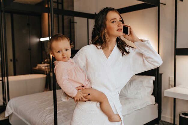 Hermosa joven madre en bata de baño blanca sosteniendo a su hija en brazos y hablando por teléfono en el dormitorio.