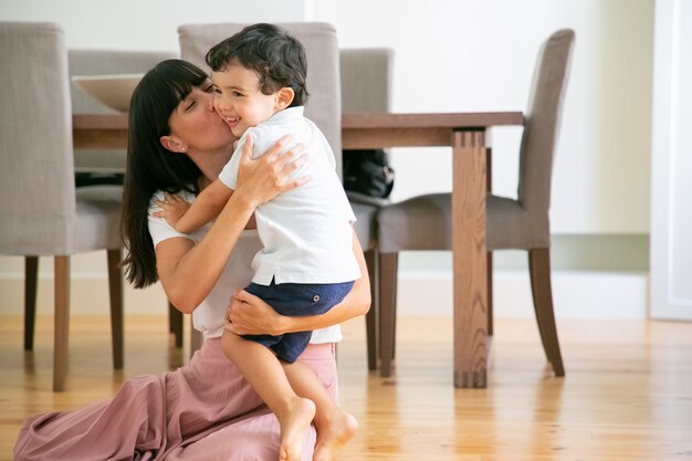 Hermosa joven madre sentada en el suelo y besando a su hijo.