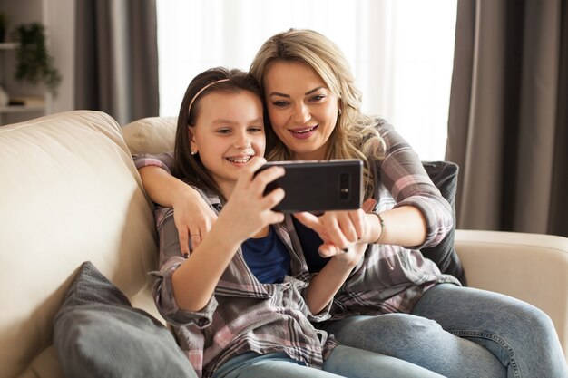 Hermosa joven madre y su hija están usando un teléfono inteligente y sonriendo sentados en el sofá de la sala de estar.