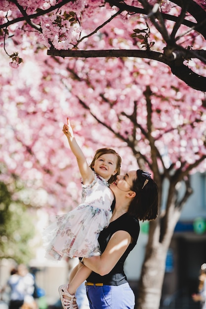 Hermosa joven madre tiene encantadora hija pequeña de pie bajo el árbol de flor rosa