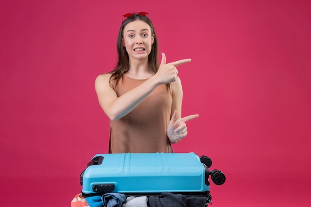 Foto gratuita hermosa joven con maleta de viaje apuntando con los dedos hacia el lado mirando confundido sobre pared rosa