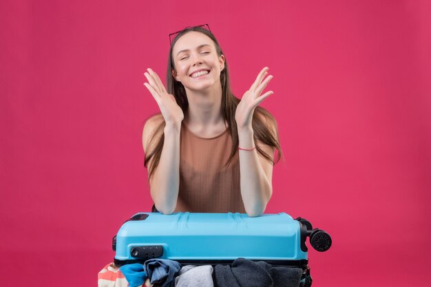 Foto gratuita hermosa joven con maleta de viaje con los ojos cerrados, sonriendo y preguntándose sobre la pared rosa