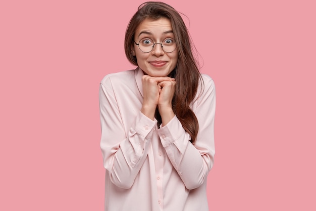 Hermosa joven mantiene las manos debajo de la barbilla, tiene los ojos llenos de felicidad, cabello largo y oscuro, vestida con una camisa elegante