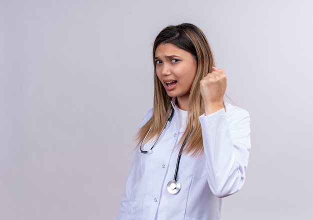 Foto gratuita hermosa joven médico vistiendo bata blanca con estetoscopio apretando el puño con cara enojada