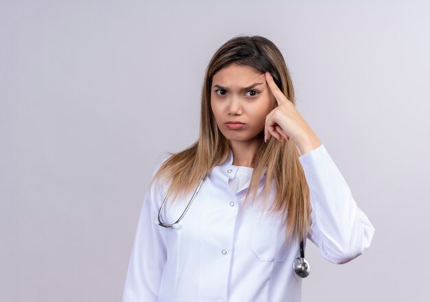 Hermosa joven médico vistiendo bata blanca con estetoscopio apuntando a la sien con el dedo concentrándose en una idea con expresión seria