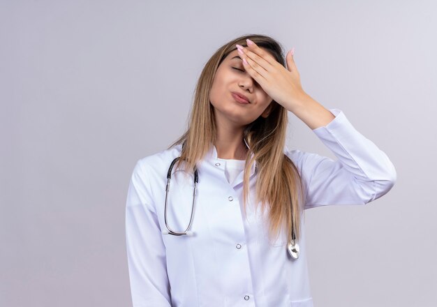 Hermosa joven médico vistiendo bata blanca con estetoscopio con aspecto cansado y con exceso de trabajo cubriendo los ojos con la mano