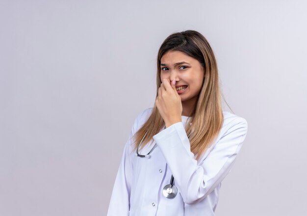 Hermosa joven médico vistiendo bata blanca con estetoscopio cerrando la nariz con los dedos sintiendo un aroma agradable