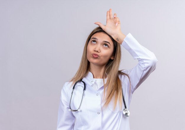 Hermosa joven médico vistiendo bata blanca con estetoscopio gesticulando más suelto cantar con las manos sobre la cabeza