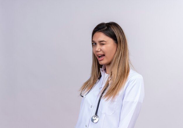 Hermosa joven médico vistiendo bata blanca con estetoscopio guiñando un ojo y sonriendo