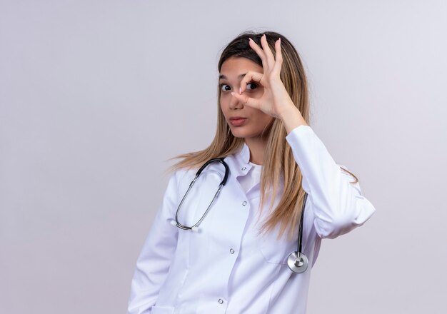 Hermosa joven médico vistiendo bata blanca con estetoscopio haciendo bien firmar con los dedos mirando a través de este signo