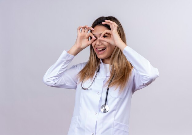 Hermosa joven médico vistiendo bata blanca con estetoscopio haciendo bien los signos con los dedos como binoculares mirando a través de los dedos sacando la lengua