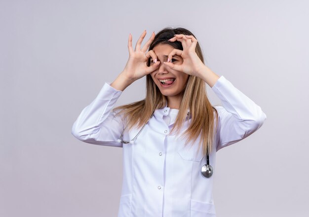 Hermosa joven médico vistiendo bata blanca con estetoscopio haciendo bien los signos con los dedos como binoculares mirando a través de los dedos sacando la lengua