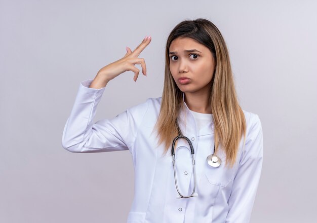 Hermosa joven médico vistiendo bata blanca con estetoscopio haciendo gesto de pistola o pistola cerca de la cara ceñuda del templo