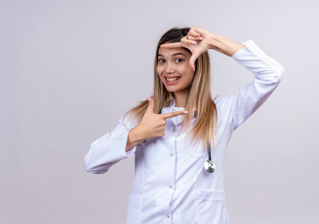 Hermosa joven médico vistiendo bata blanca con estetoscopio haciendo marco con los dedos sonriendo mirando a través de este marco