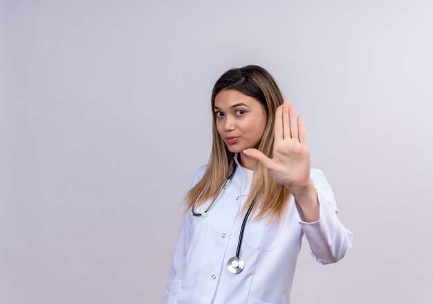 Hermosa joven médico vistiendo bata blanca con estetoscopio haciendo señal de stop con la mano abierta con cara seria con expresión seria