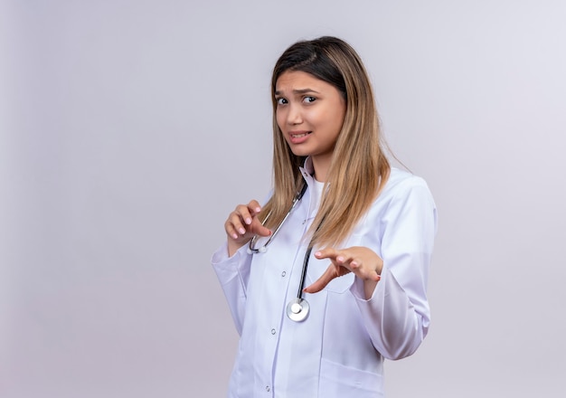 Hermosa joven médico vistiendo bata blanca con estetoscopio levantando las palmas en gesto de rechazo con expresión de disgusto
