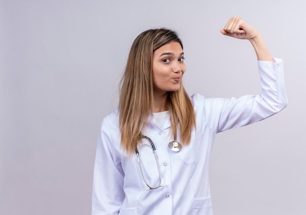 Hermosa joven médico vistiendo bata blanca con estetoscopio levantando el puño mostrando bíceps como un ganador sonriendo confiado