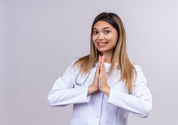 Hermosa joven médico vistiendo bata blanca con estetoscopio mano en oración gesto de namaste sintiéndose agradecido y feliz