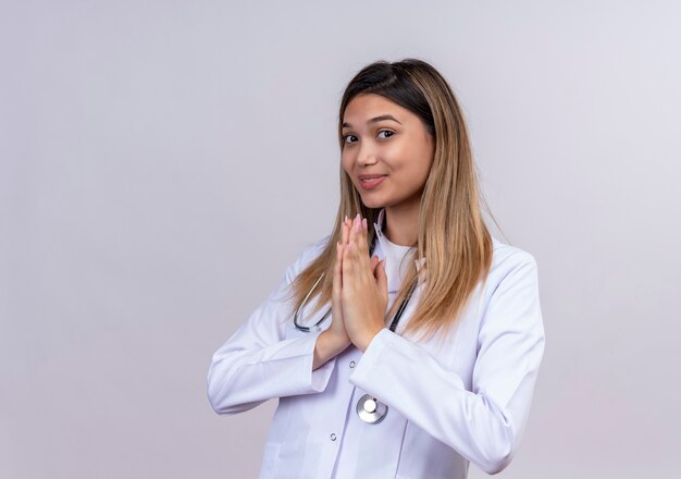 Hermosa joven médico vistiendo bata blanca con estetoscopio mano en oración gesto de namaste sintiéndose agradecido y feliz