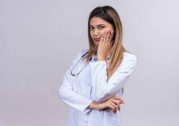 Hermosa joven médico vistiendo bata blanca con estetoscopio mirando aburrido con la mano en la barbilla esperando