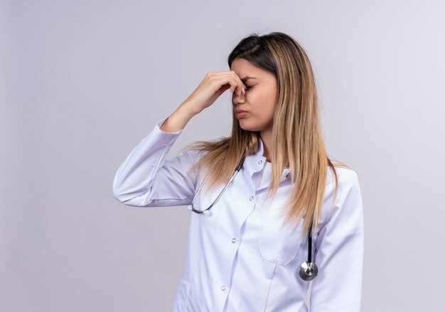 Hermosa joven médico vistiendo bata blanca con estetoscopio mirando cansado y aburrido tocando la nariz entre los ojos cerrados