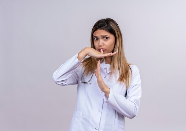 Hermosa joven médico vistiendo bata blanca con estetoscopio mirando con el ceño fruncido y haciendo gesto de tiempo con las manos