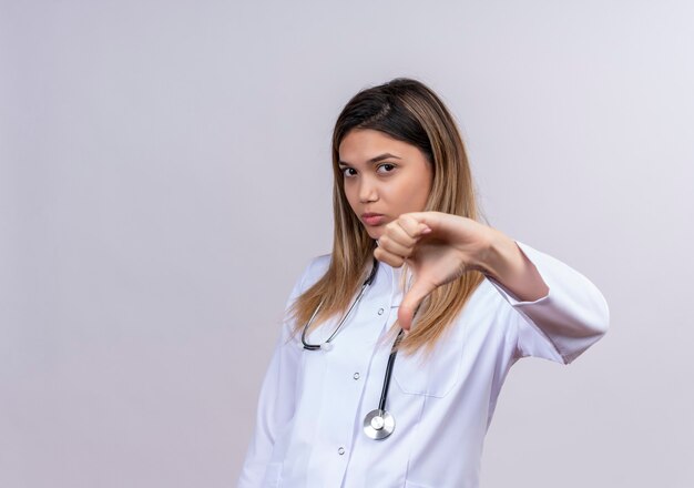 Hermosa joven médico vistiendo bata blanca con estetoscopio mirando con el ceño fruncido mostrando disgusto