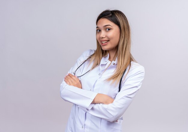 Hermosa joven médico vistiendo bata blanca con estetoscopio mirando confiado de pie con los brazos cruzados.