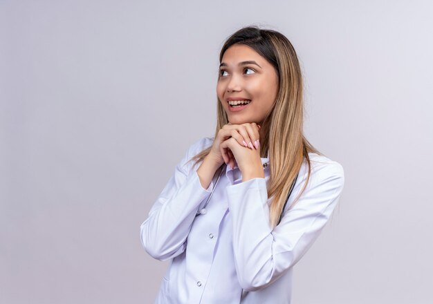 Hermosa joven médico vistiendo bata blanca con estetoscopio mirando a un lado cogidos de la mano juntos salieron esperando sorpresa