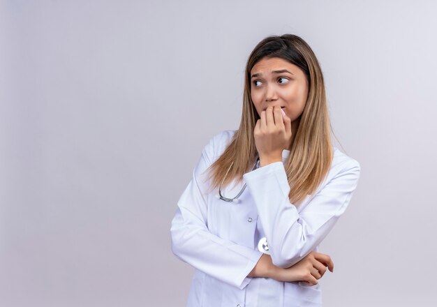 Hermosa joven médico vistiendo bata blanca con estetoscopio mirando a un lado estresado y nervioso morderse las uñas