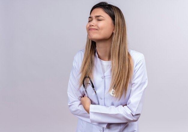 Hermosa joven médico vistiendo bata blanca con estetoscopio mirando mal tocando su vientre que sufre de dolor