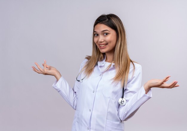 Hermosa joven médico vistiendo bata blanca con estetoscopio mirando positivo y feliz sonriendo extendiendo las palmas hacia los lados